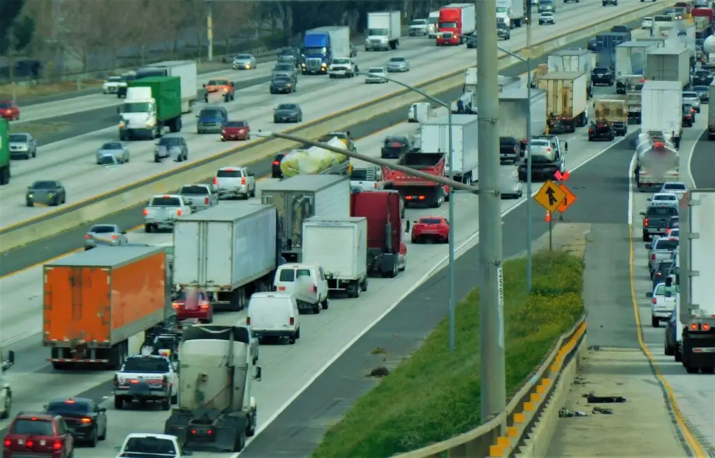 Busy multi-lane highway with numerous trucks and cars in heavy traffic, concrete barriers, and a grassy median; construction cones and equipment visible on the shoulder.