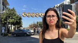 AustinDomainAccident | V.Valenzuela Law | V.Valenzuela Law A young woman takes a selfie on a sunny street; behind her a car crash blocks the road under an arch sign reading INDOMA with bystanders nearby.